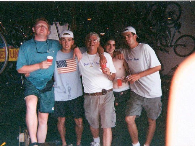 Five people in casual wear pose with drinks in a garage with bicycles in the background.