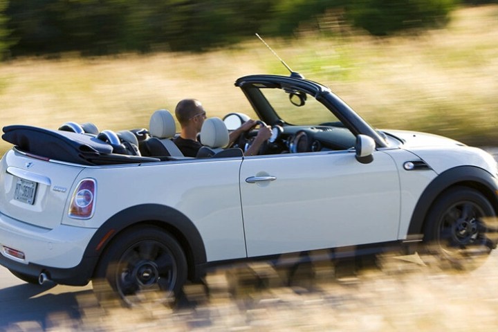Man driving a white convertible Mini Cooper on a sunny road with blurred grass in the background.