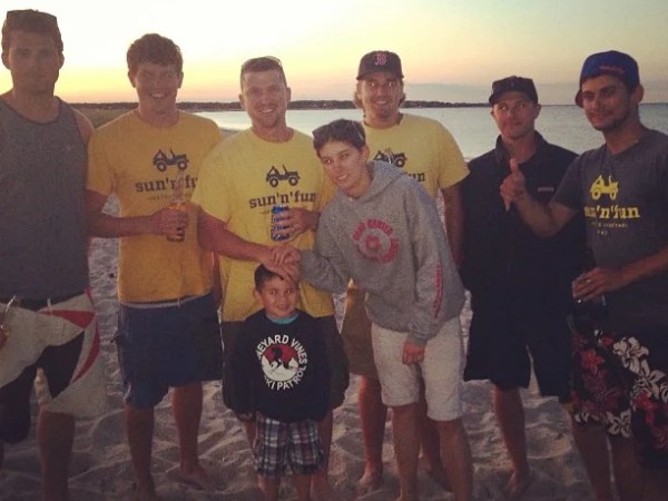 Group of seven men and a child posing on a beach at sunset, some wearing yellow 'sun n' fun' shirts.
