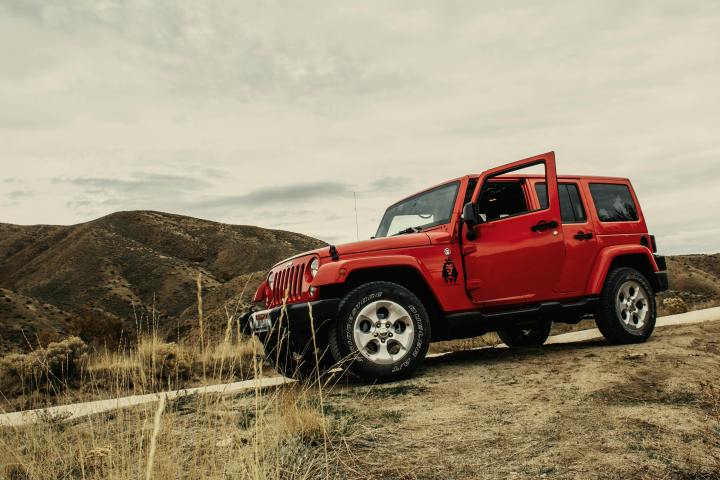 Red SUV with open door on a dirt road near hills under a cloudy sky.