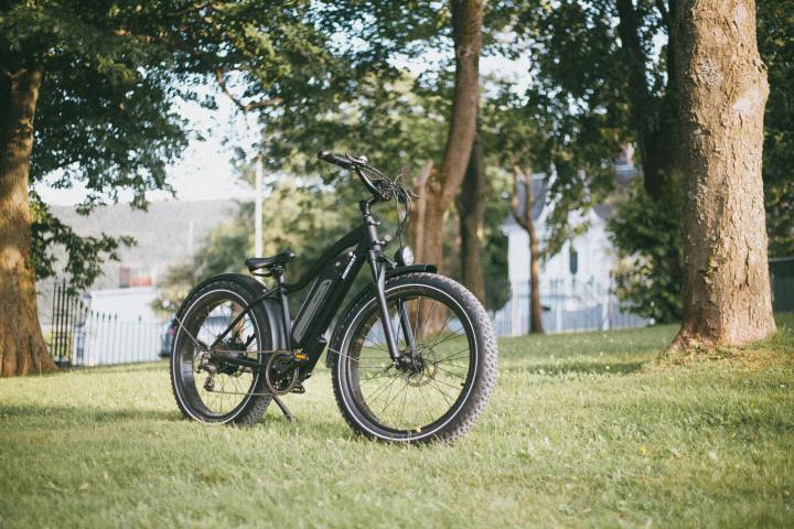 Black electric bike on grassy area surrounded by trees and distant buildings.